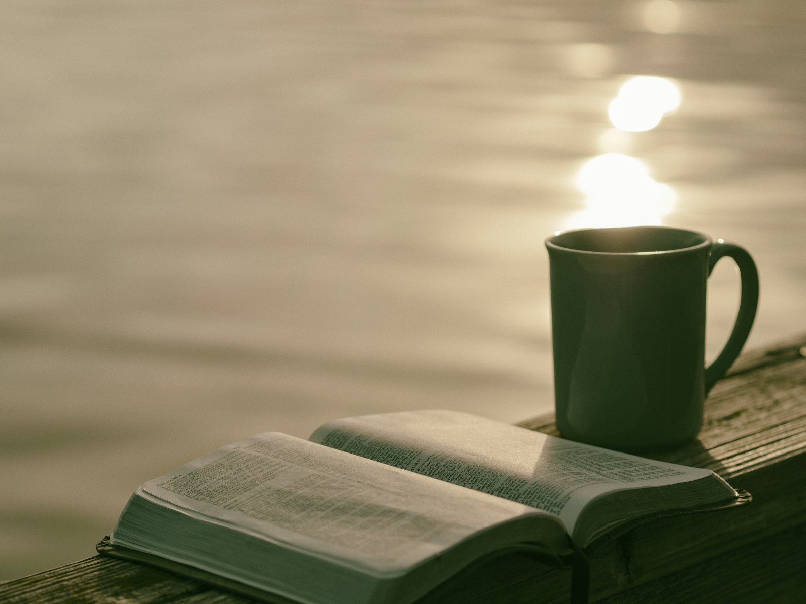 An open Bible and a mug resting on a wooden railing beside calm water at sunset