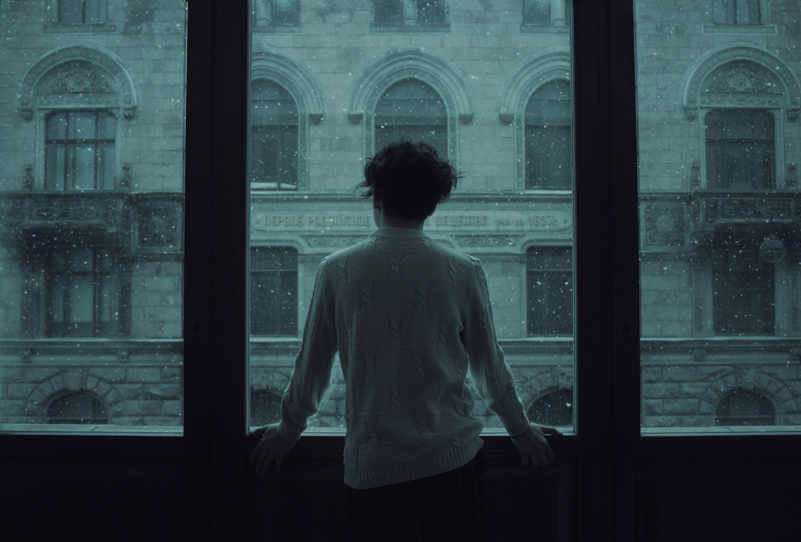 A person standing alone facing a tall rain-streaked window overlooking an old city building