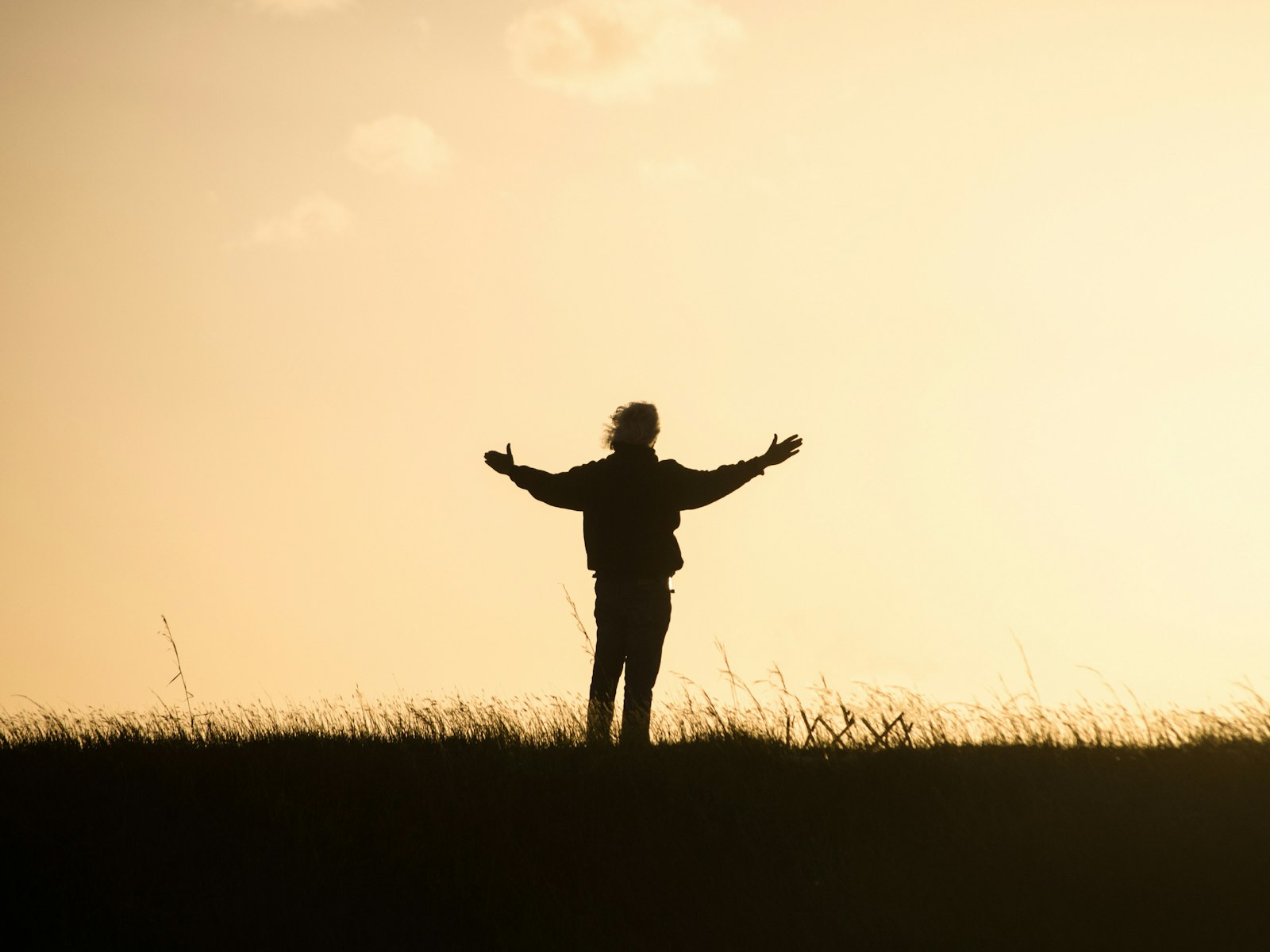 Silhouette of a person standing in a field with arms open wide toward a golden sunset sky