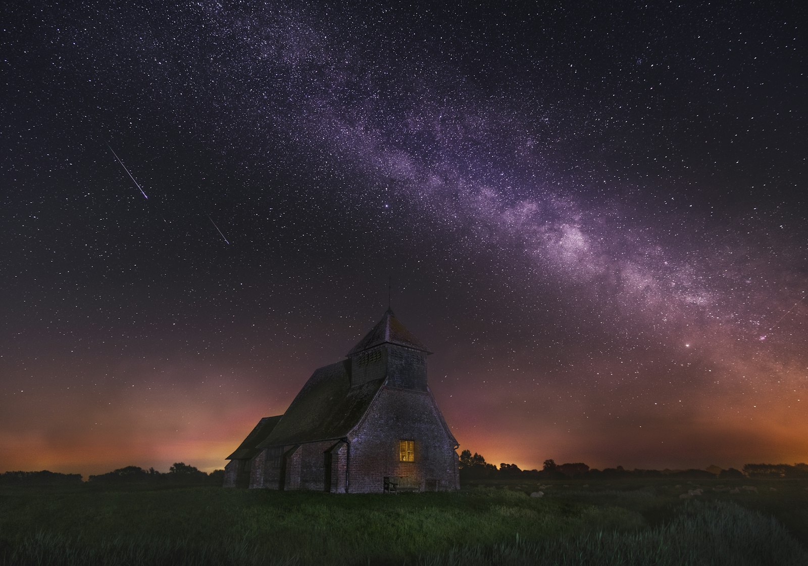 A small stone church glowing with light beneath a star-filled night sky and the Milky Way