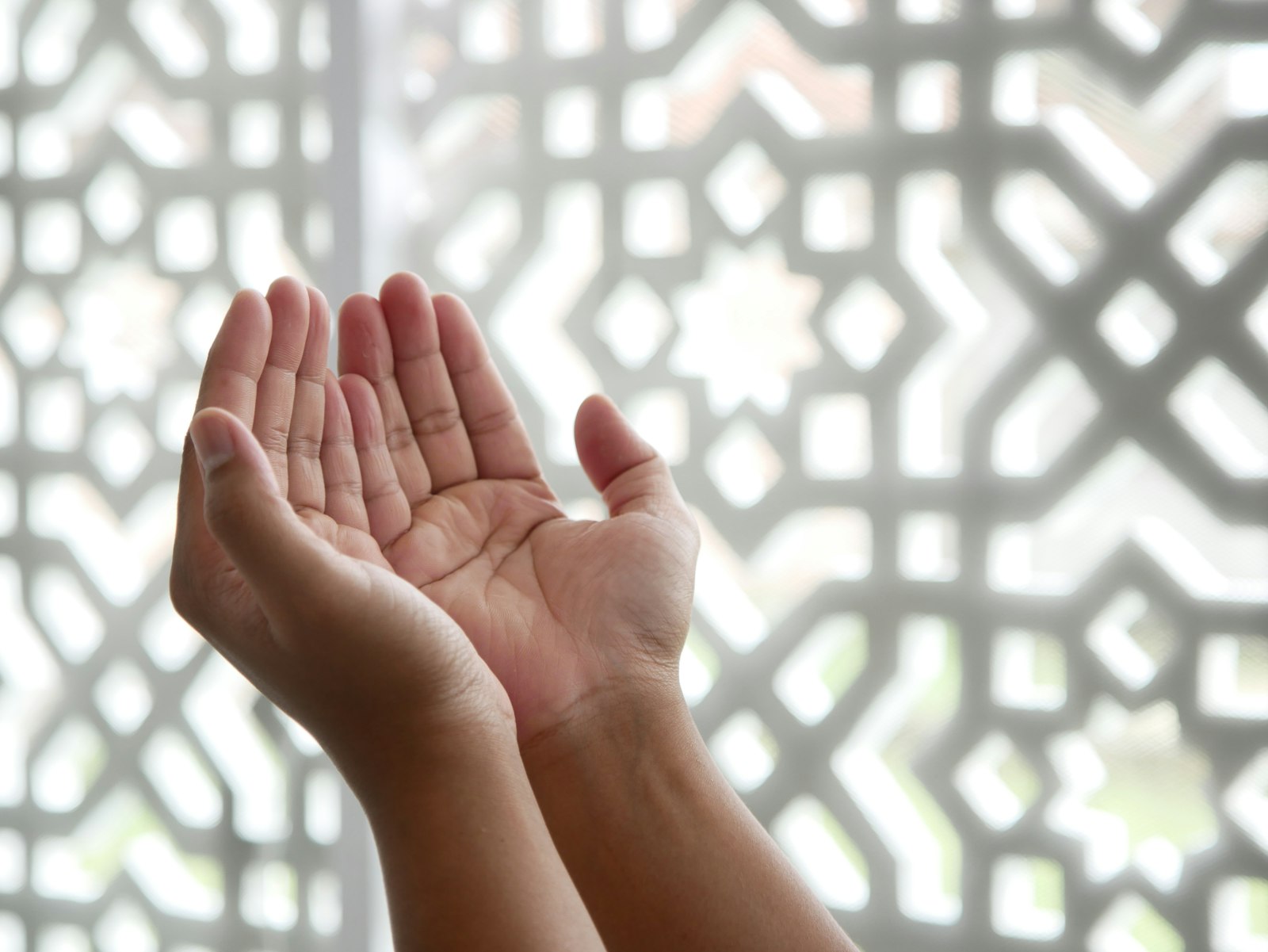 Open hands raised in prayer, lit by soft patterned light from a window